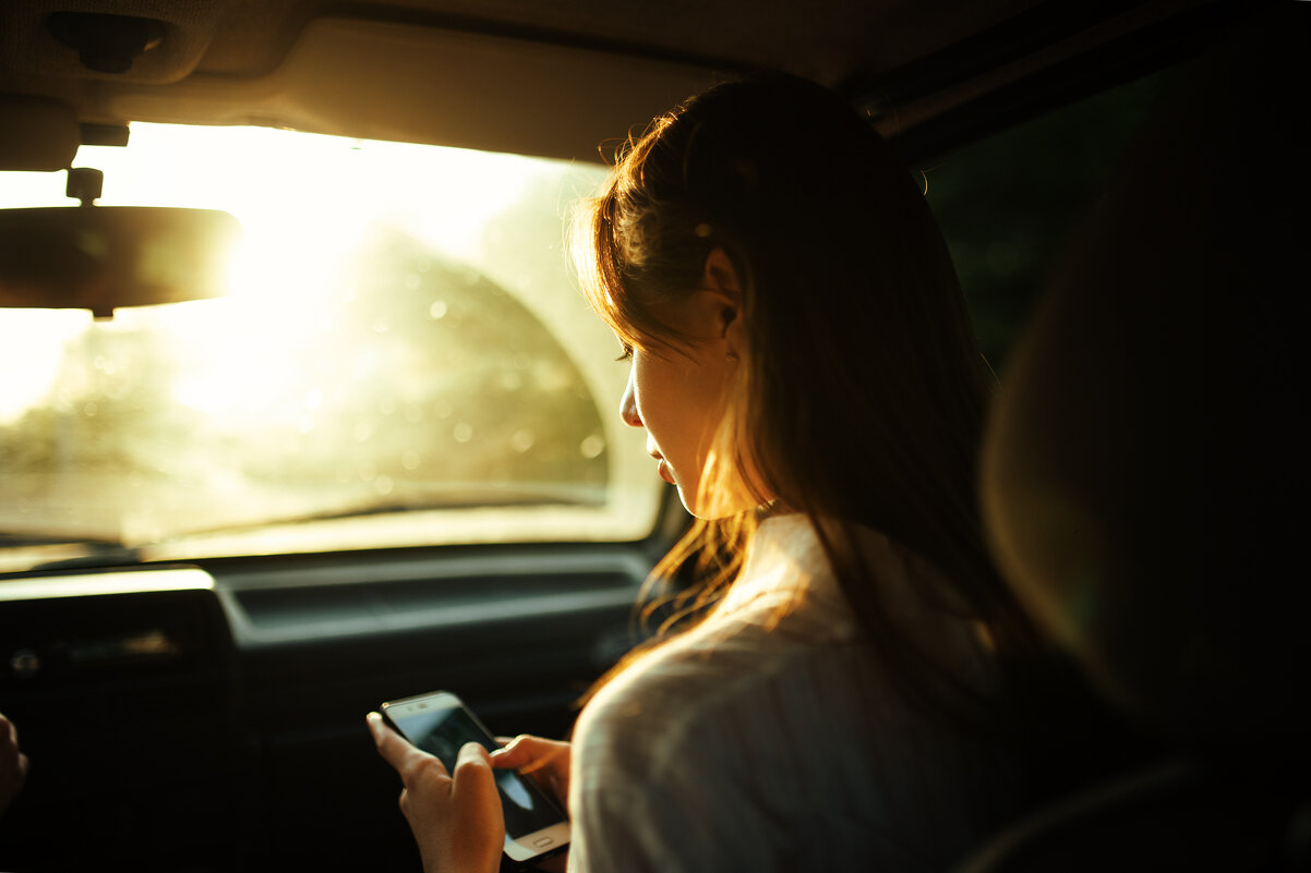 Девушка держит телефон в руке сидя в машине на фоне солнечного блика во время заката - Lenar Abdrakhmanov